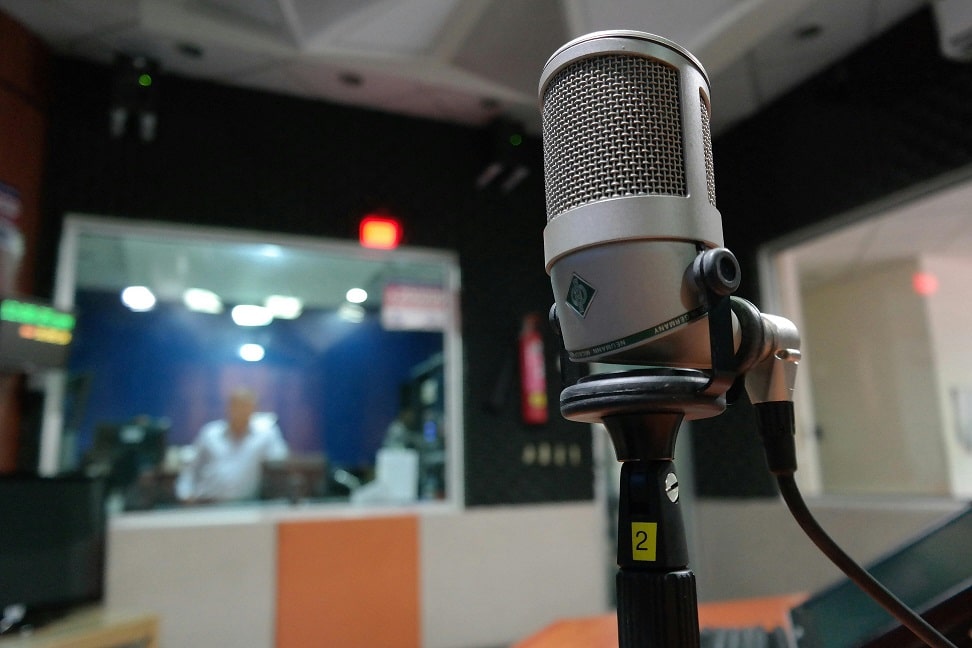 A microphone sit on a mic stand in a recording studio. In the background, a blurred image of a man can be seen through the studio glass.