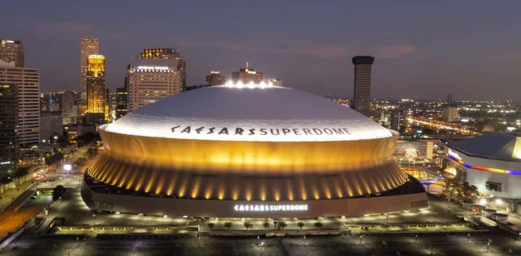 An exterior view of the Caesar's Superdome in New Orleans at night. The base of the stadium is lit in yellow lights.