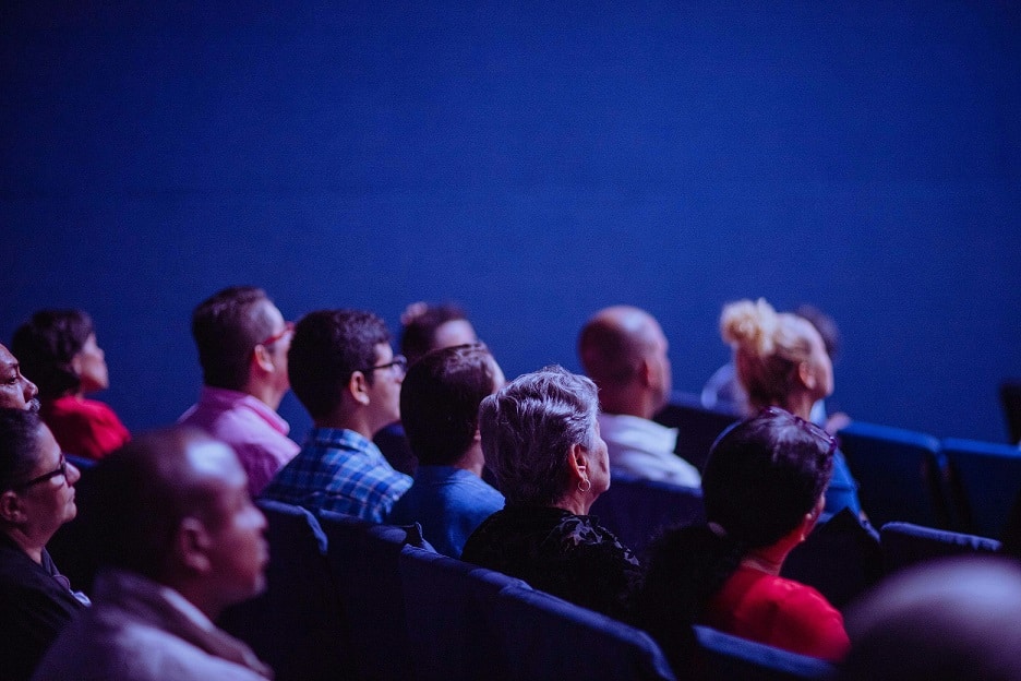 Viewed from behind, rows of people sitting in stadium seats watching a presentation