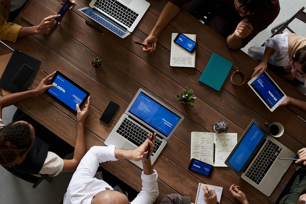 Viewed from above, people work at a desk using laptops, cell phones, and tablets.