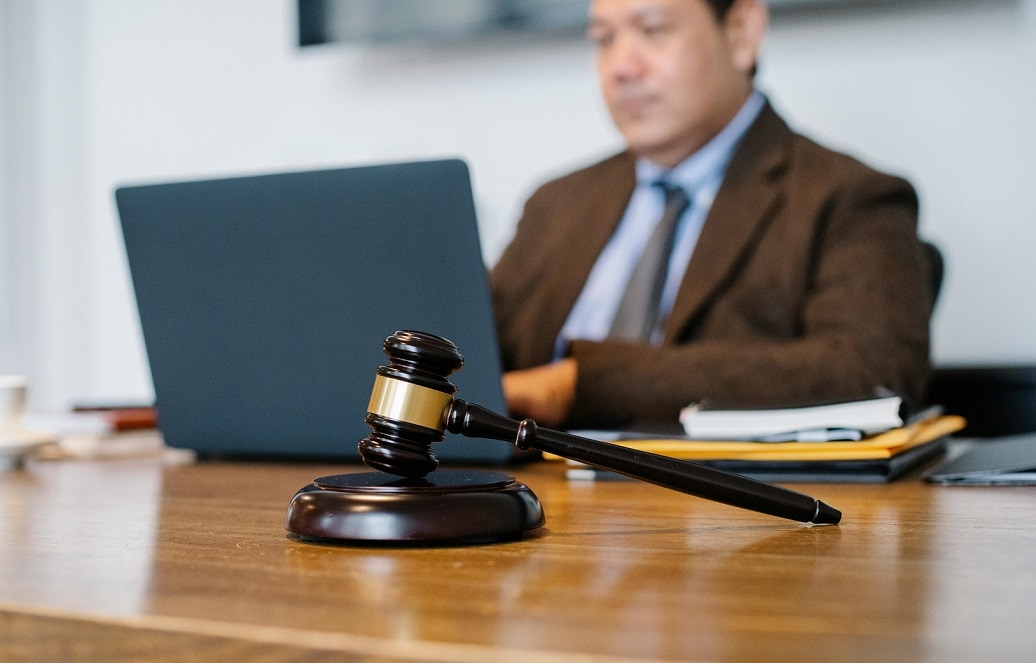 A gavel rests on a desk in the foreground while a man types at a laptop in the background