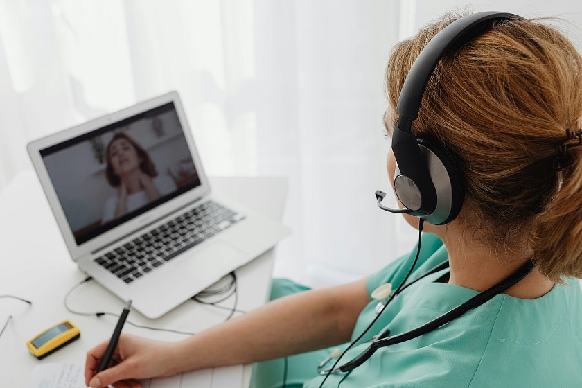 Seen from over-the-shoulder, a doctor wearing a headset speaks to a patient via a conference call on a laptop. The patient is pointing to her throat.