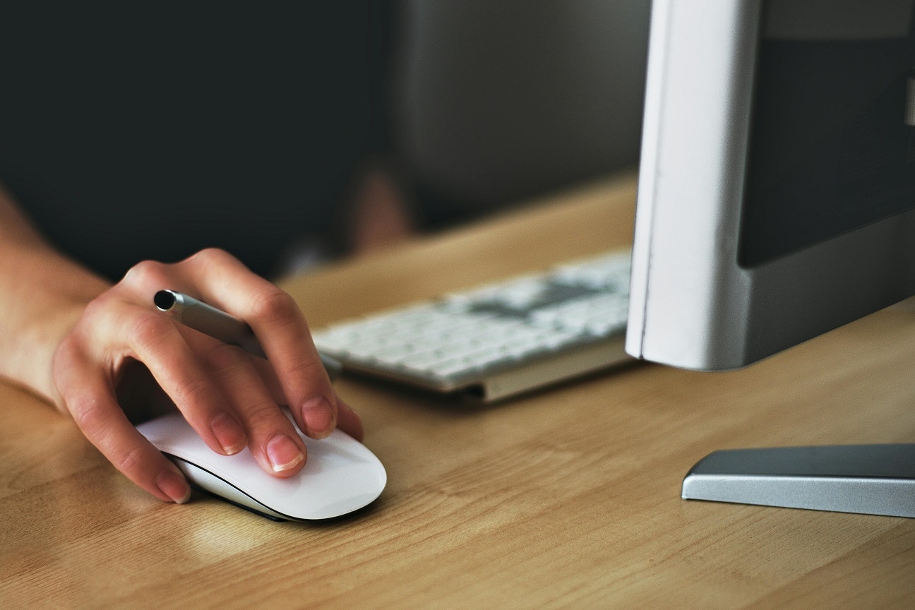 A woman's hand moves a mouse atop a desk. A keyboard and monitor are seen.