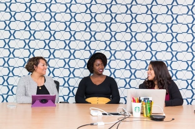 Three women sit and takl at a long conference table situated against a blue and white background.