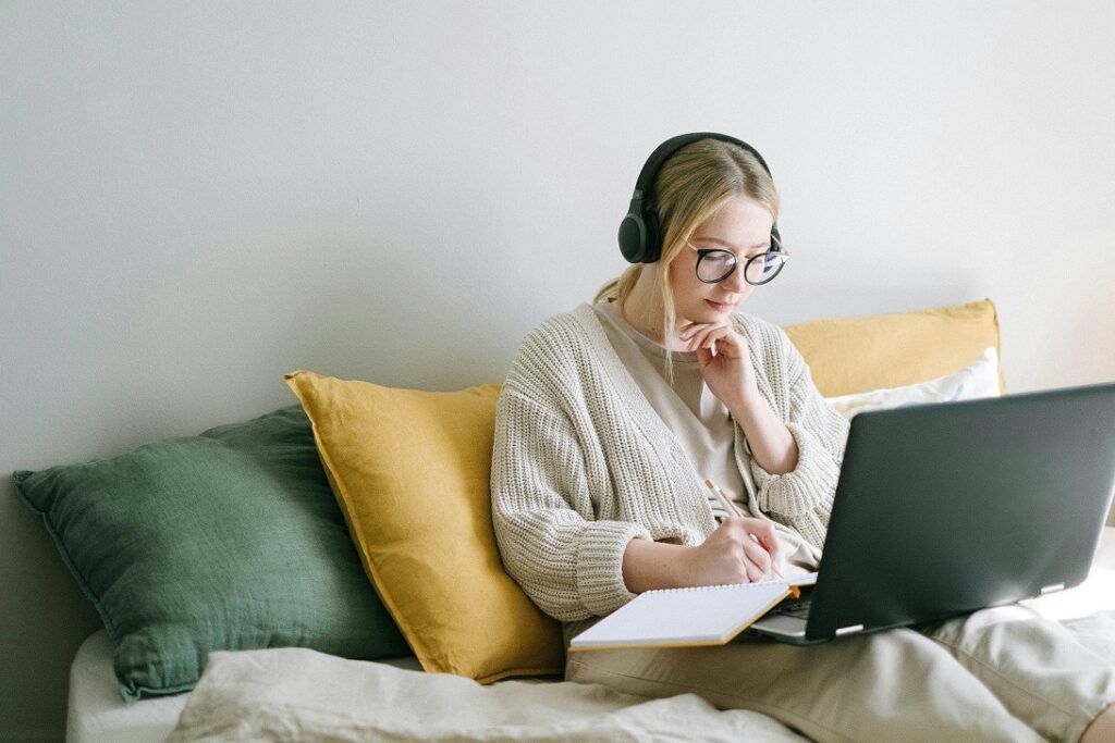 A woman with blonde hair and glasses sits on a couch. She is wearing headphones as she works on her laptop
