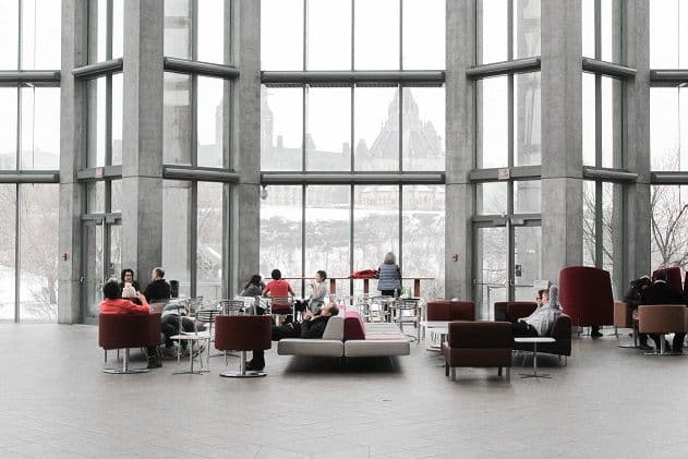 Wide view of employees sitting in a bright white office lounge with tall horizontal windows along the back wall.