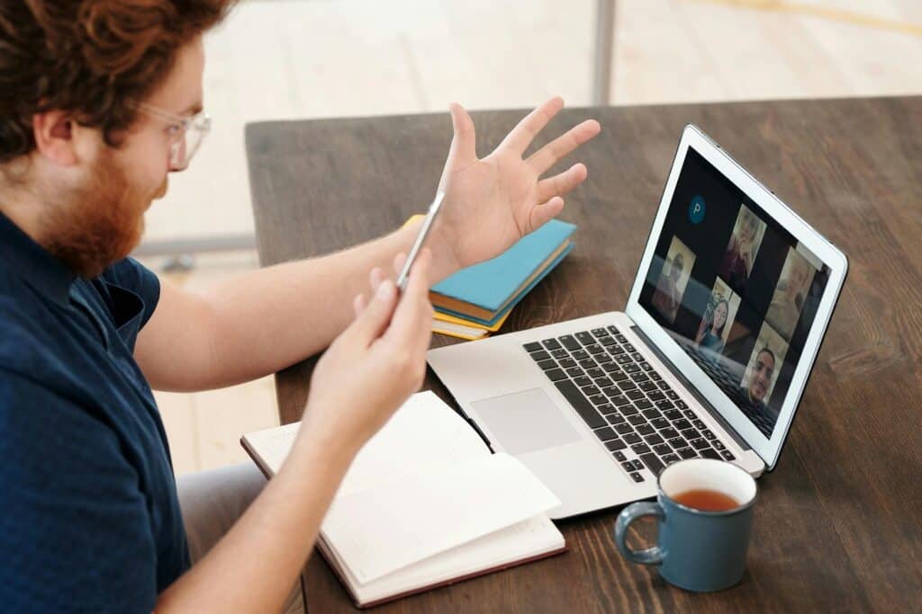 A man sits at a desk and interacts with a virtual meeting on his laptop.
