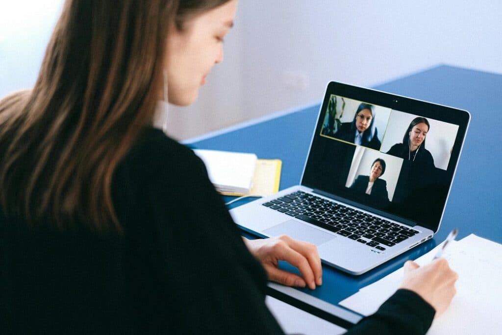 A woman seated at a desk looks at her laptop screen and speaks with three people during a virtual meeting.