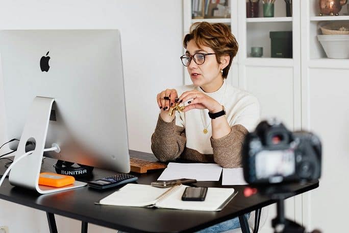 A woman with short reddish hair, wearing glasses, sits in front of a computer screen.