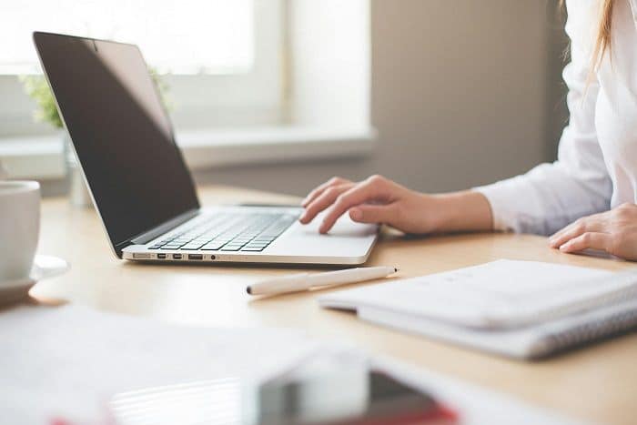 A woman's hand rests on a laptop keyboard. In the foreground, on a desk, is a pencil and notebook.