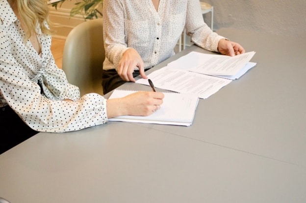 Two women work on a document at a desk, only their hands are visible as they point out items on papers.
