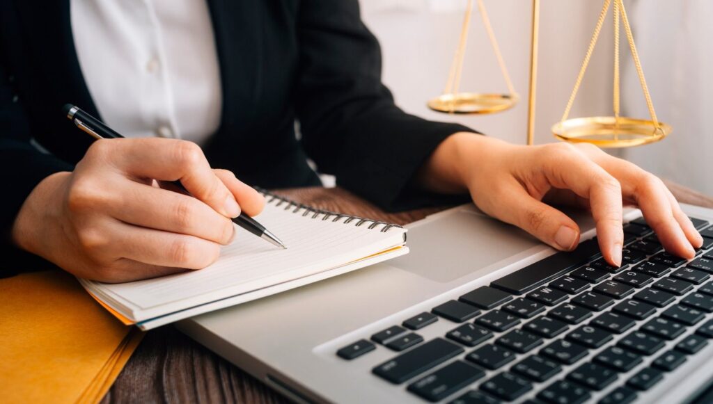 Close up of a woman's hands as she uses a computer keyboard. In her right hand is a pen as she writes on a tablet