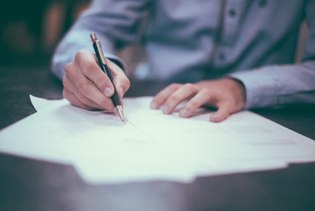 A man's hands are seen working on a document at his desk. He holds a pen in his right hand.