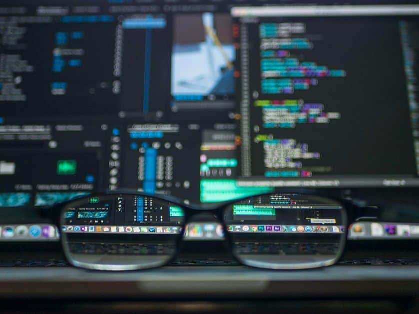 A pair of eyeglasses sits on a table in front of a computer screen. The screen can be seen magnified through the glass lenses.