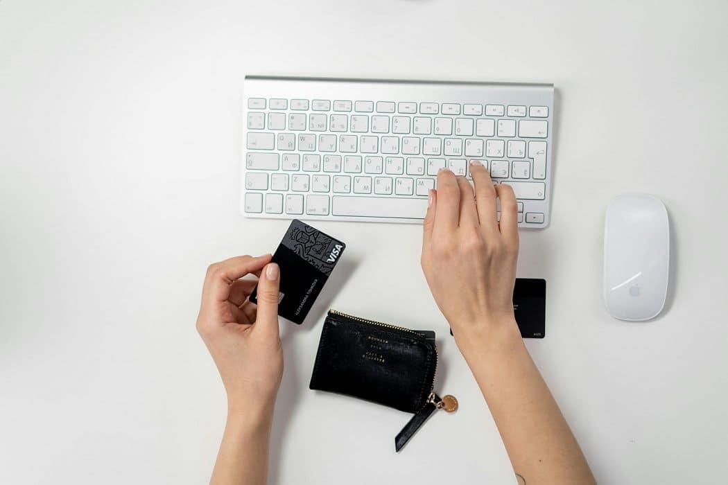A person hands on a stark white keyboard. The right hand rests on the keyboard, the left is holding a black VISA card.