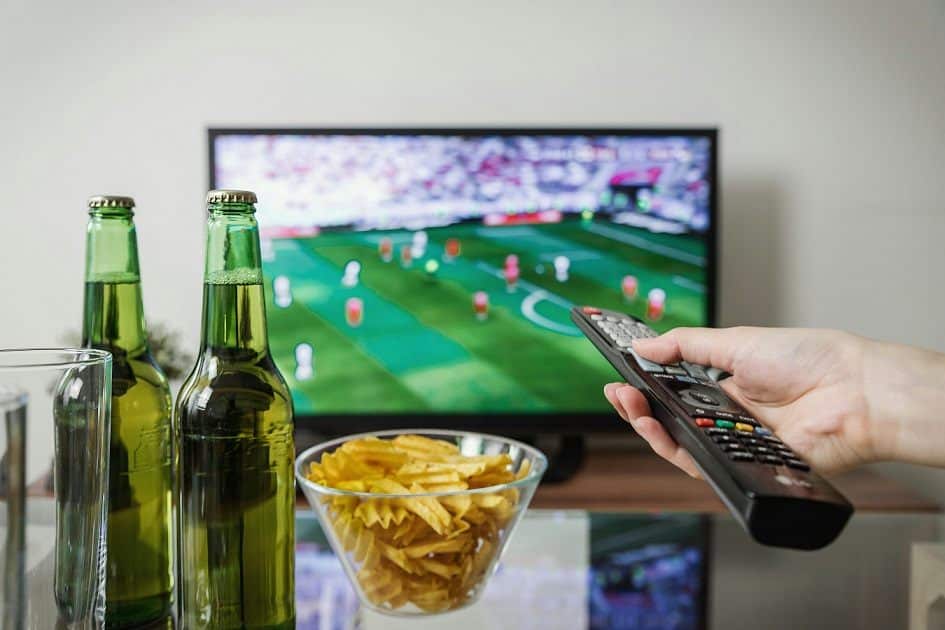 A person points a remote control towards the television. A soccer game can be seen on the screen, a bowl of chips and two bottles of beer sit on a table in front.