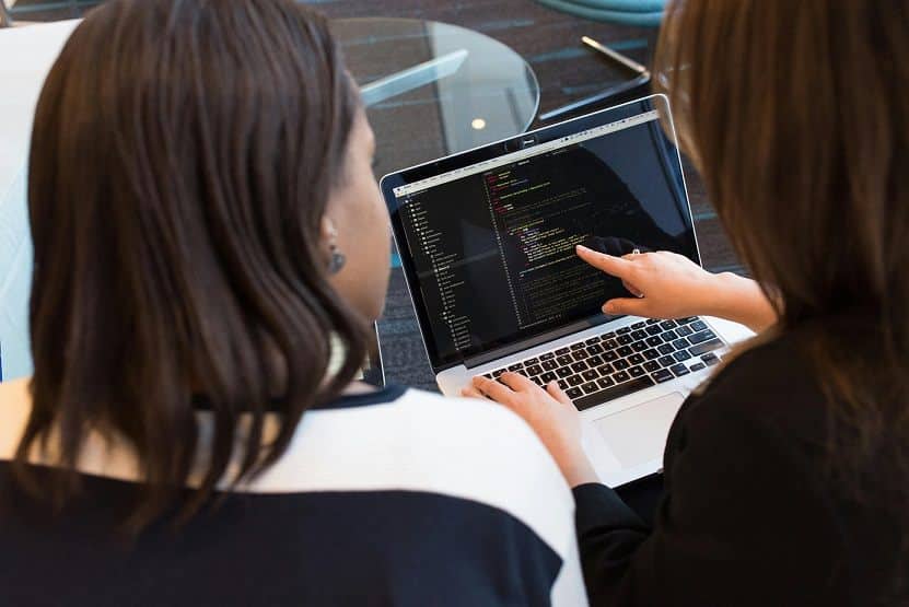 Two women look at a laptop computer screen, one is pointing to a line of text on the screen.