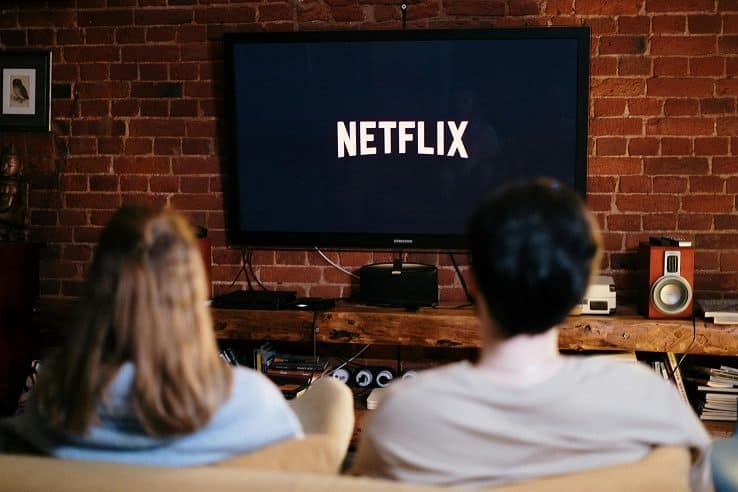 A man and a woman, viewed from behind, sit on a couch watching television. On the TV screen is the Netflix logo