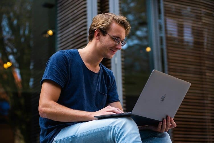 A man with brown hair and wearing a blue shirt sits outside and works on his laptop