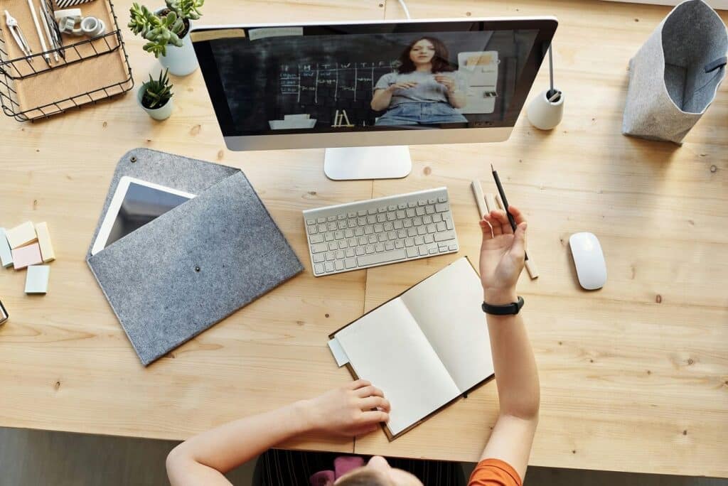 A bird's-eye view of a young woman sitting in front of a computer monitor taking a remote learning course. A teacher can be seen on the screen.