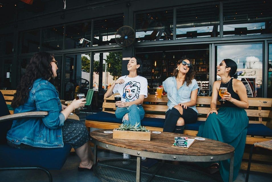 Four women sitting in an outdoor cafe, laughing and drinking beers