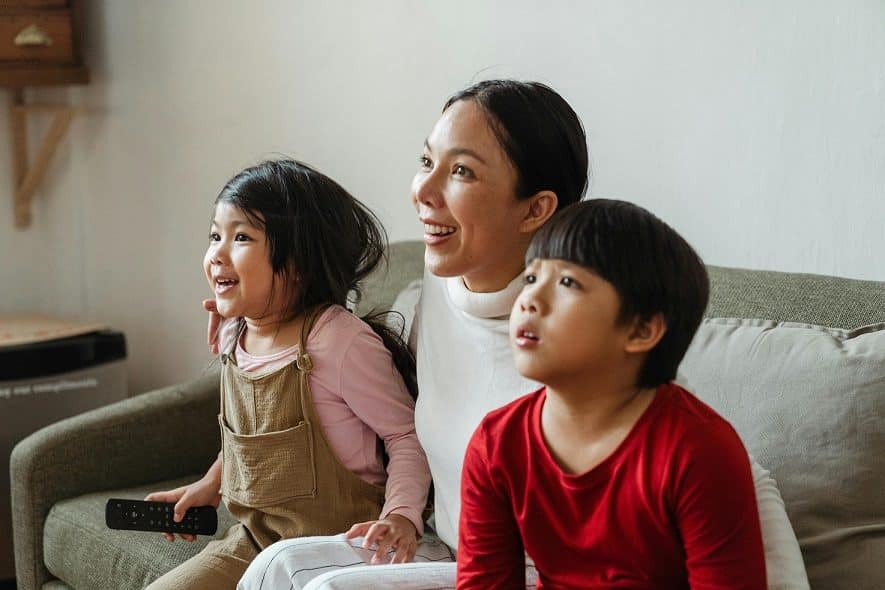 A woman with dark hair and two children -a boy and a girl - sit on a couch and watch television.
