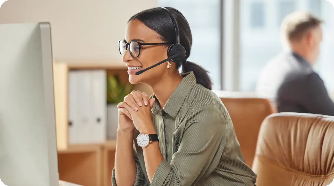 A woman with brown hair and glasses sits at a desktop with a headset on engaged in a call