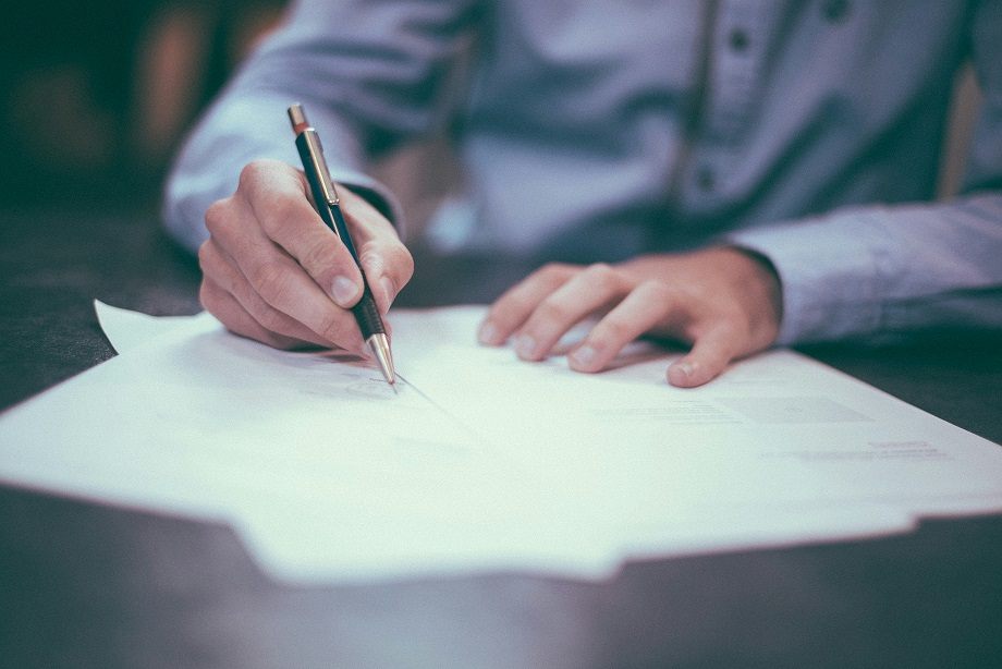 Close up of hands working on a contract. The right hand holds a pen and is writing on a white sheet of paper.