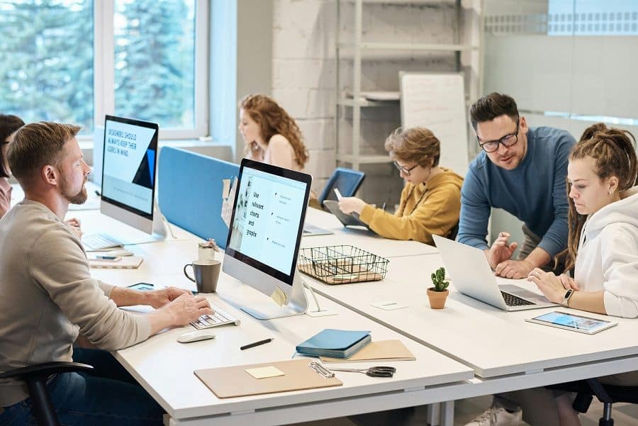 Men and women sitting at computer workstations. A man in a blue shirt with dark hair stands next to a woman with brown hair wearing a white shirt as if to be showing her something on the laptop.