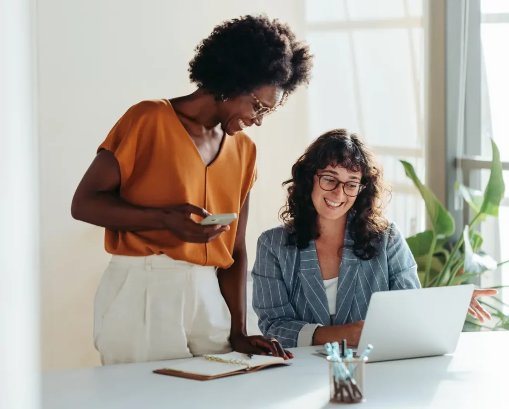 Two women hover over a laptop discussing what is on it and smiling. One stands in an orange shirt and white pants while the other sits and wears glasses and a blue shirt.