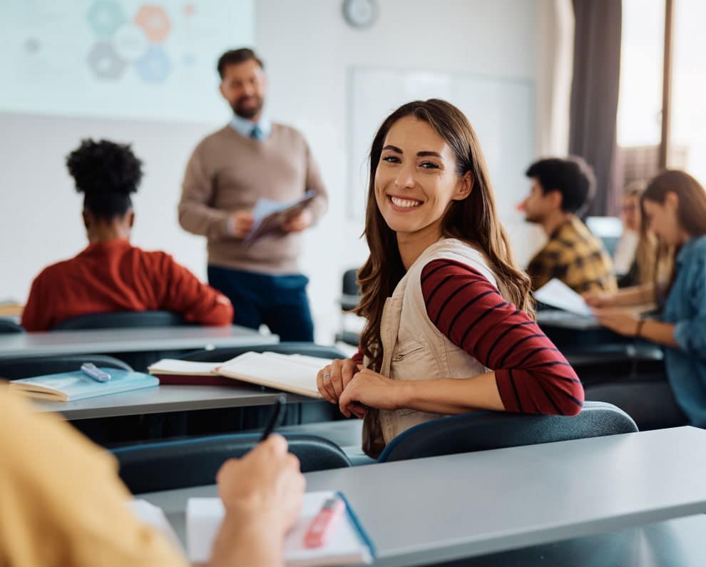 A female college student with brown hair and a striped shirt turns around to smile while in a classroom with her instructor seen standing in the background teaching and other students sitting and learning around her
