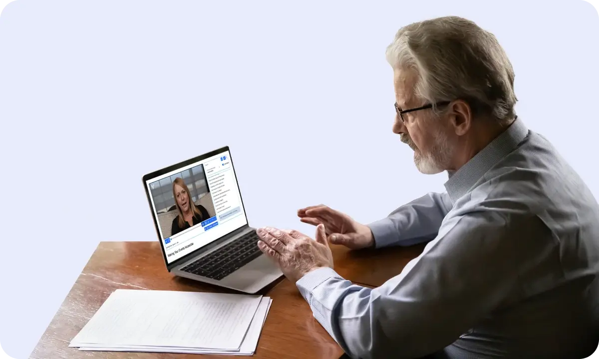An older man with grey hair sits working from a desk with a laptop watching a video