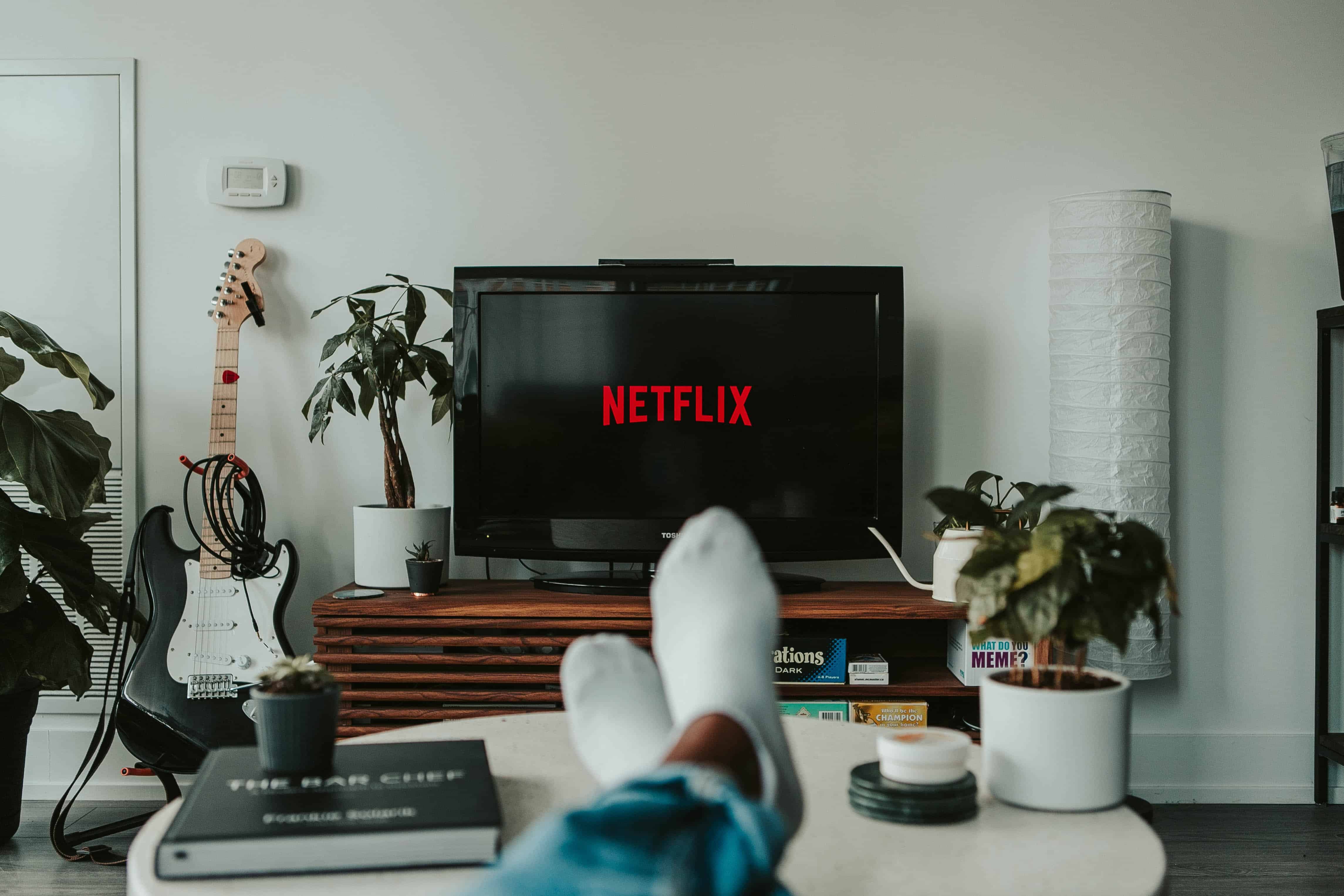 A person at home with his feet up on a coffee table with a TV visible in front of him and Netflix on the screen
