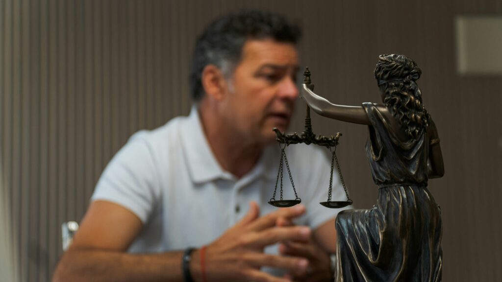 A middle aged man in a short sleeve polo is seen talking while a legal statue of a person holding a balance is seen in front of him