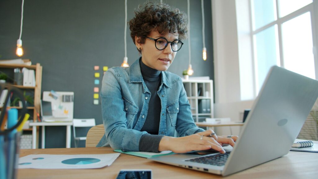 A woman with short curly brown hair and glasses wears a jean jacket and is shown in an office working from an open laptop.
