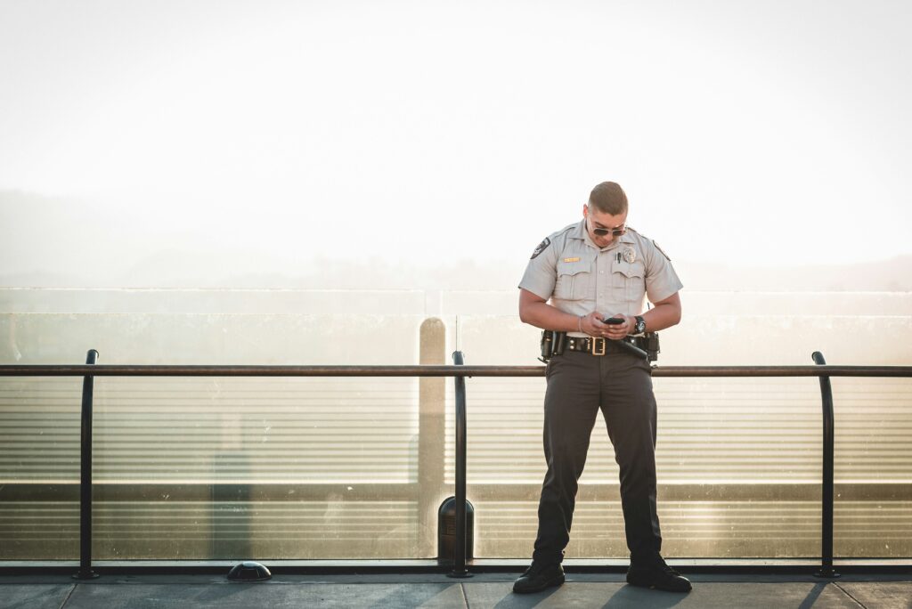 A police officer in a tan uniform is seen standing along a bridge while using his phone