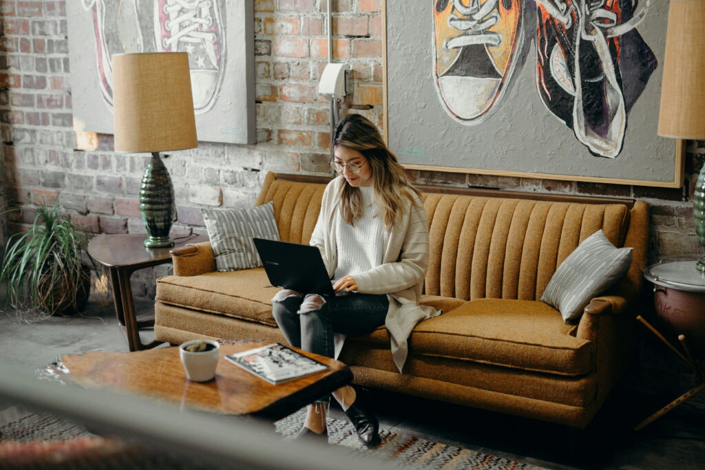 A young professional woman works from her laptop watching video from a tan couch.
