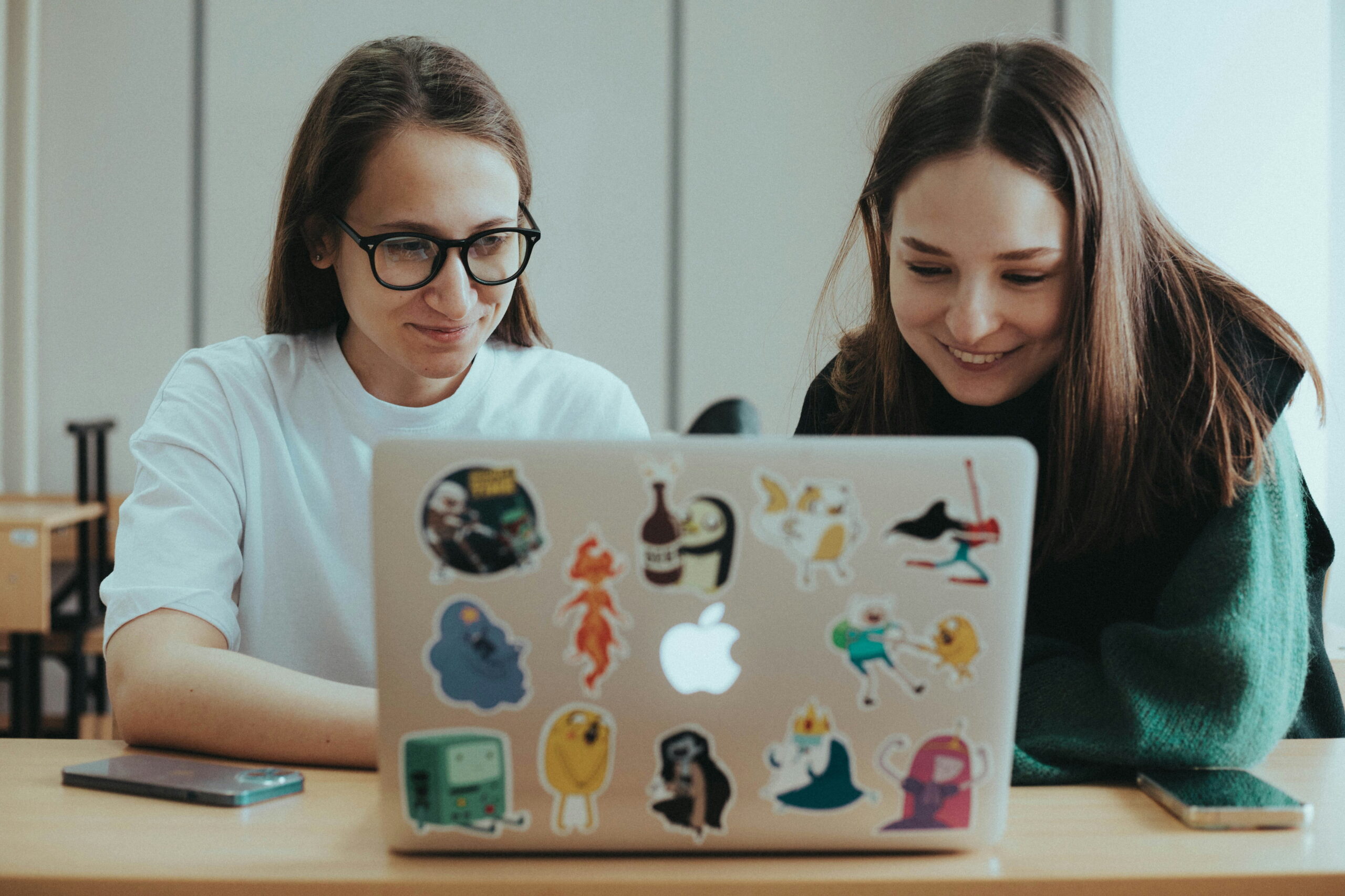 Two young women look together at the screen on a Mac laptop with stickers covering it