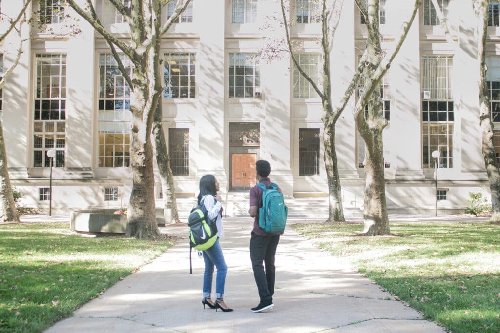 Two students - one woman and one man - stand outside on a university campus in front of a white university building talking