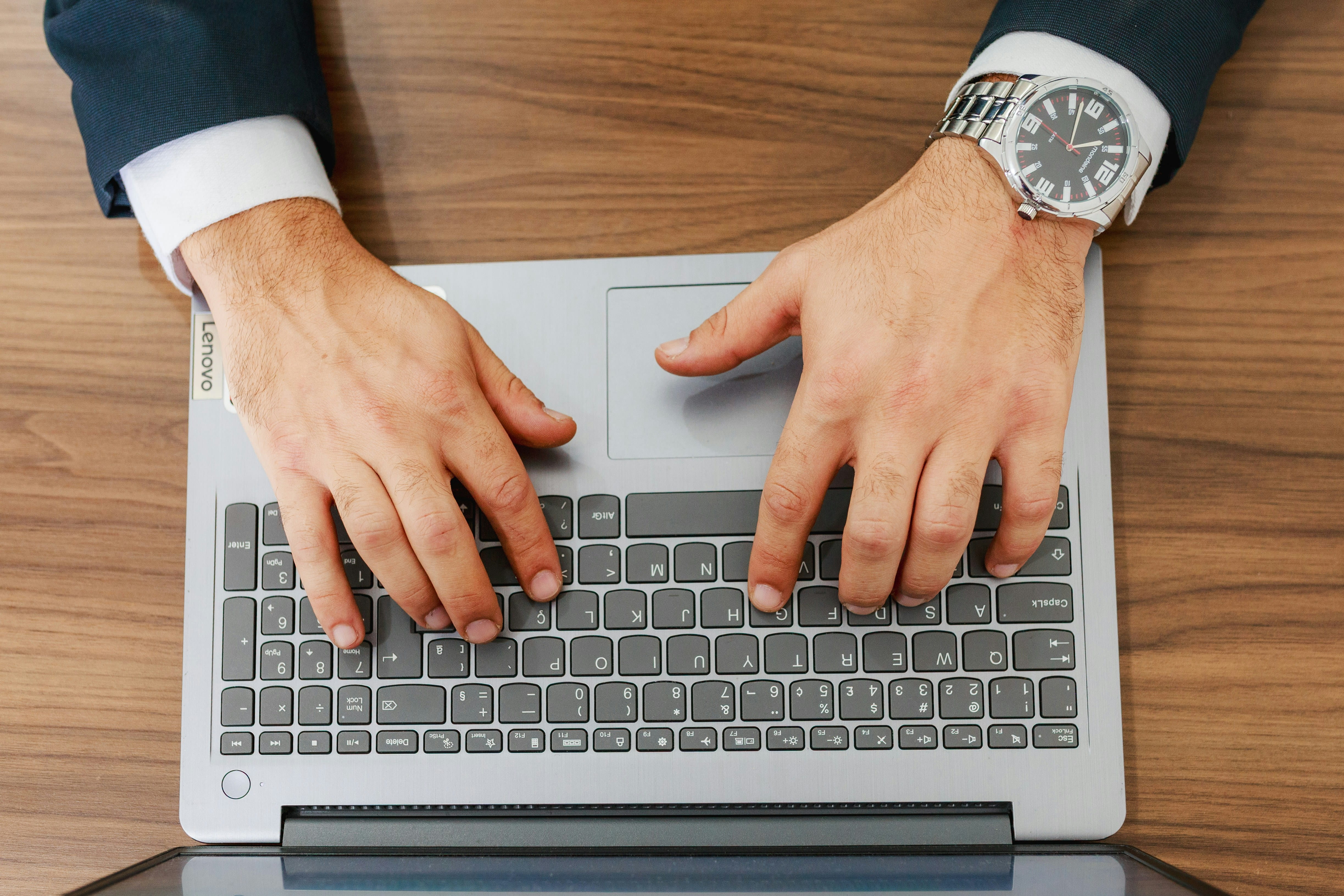 The hands of a man wearing a suit and a watch are shown typing on a Lenovo laptop