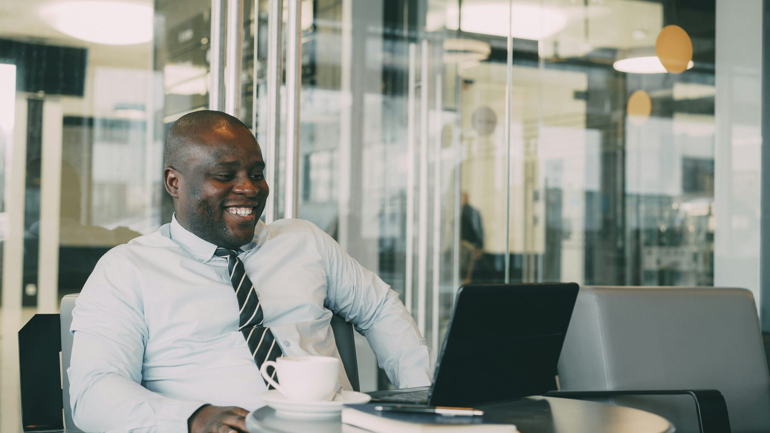 A smiling Black businessman in a light blue dress shirt and striped tie sitting at a table with a laptop and a cup of coffee in a modern office setting.