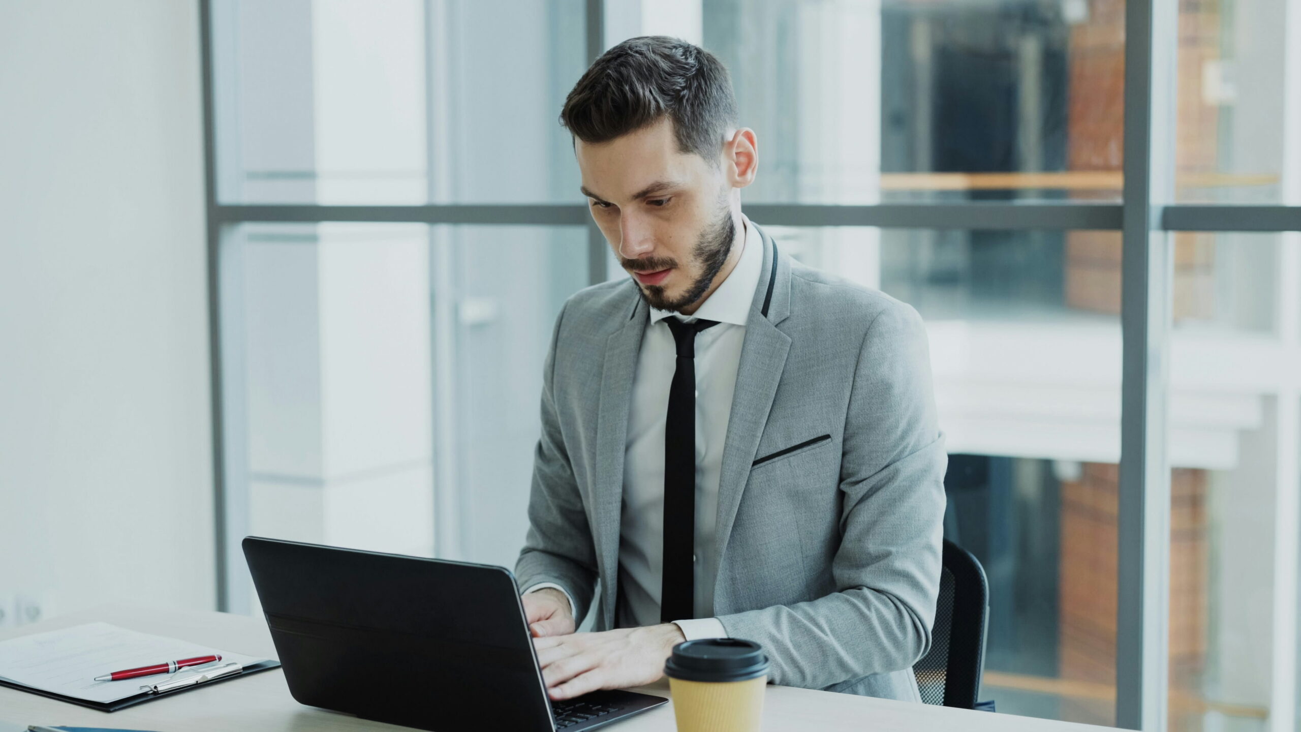 A bearded businessman wearing a grey suit jacket, white shirt, and black tie focused on typing on a laptop at a desk next to a to-go coffee cup.