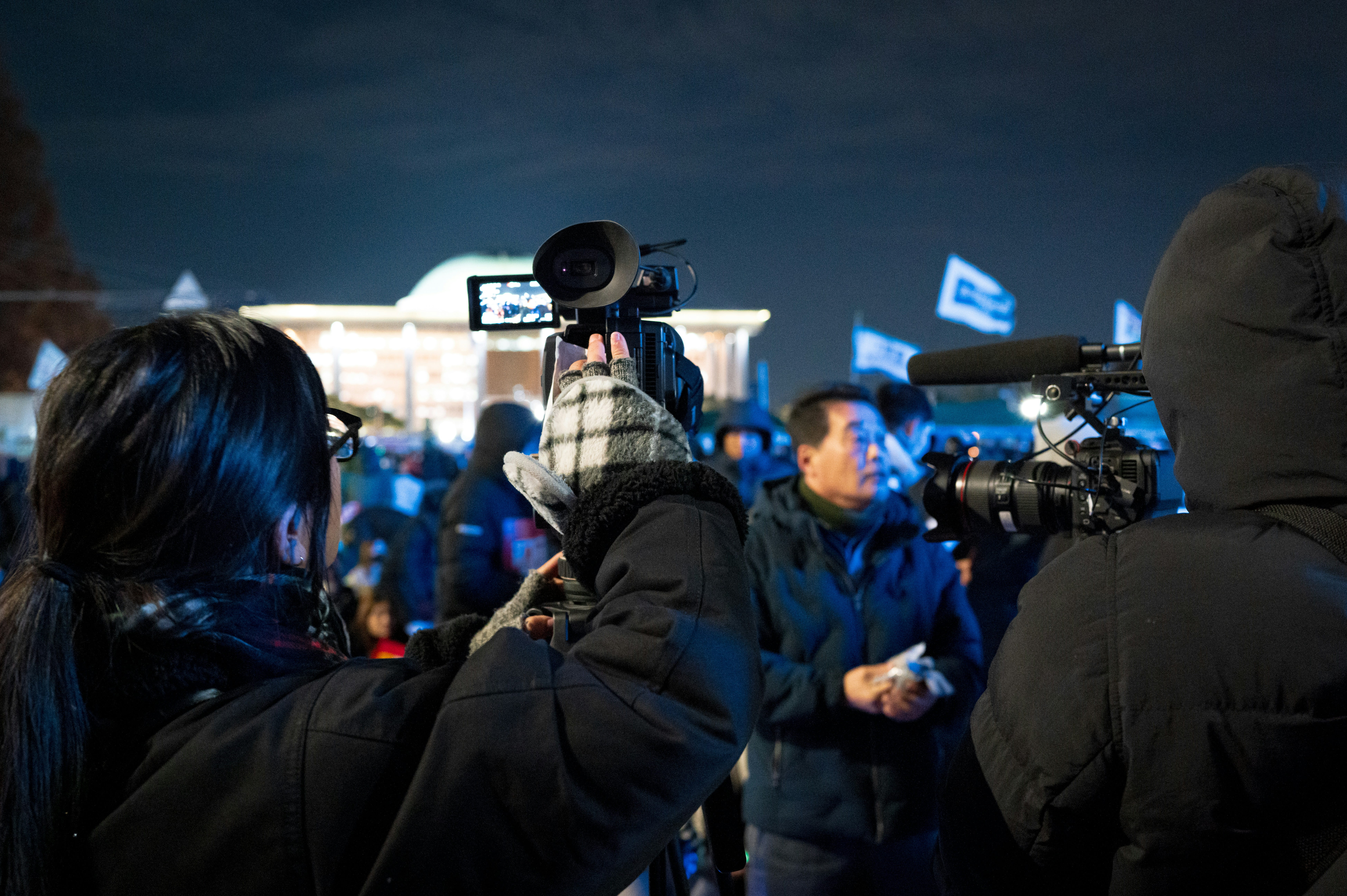 Cameramen recording an outdoor gathering or public event at night under dark skies.