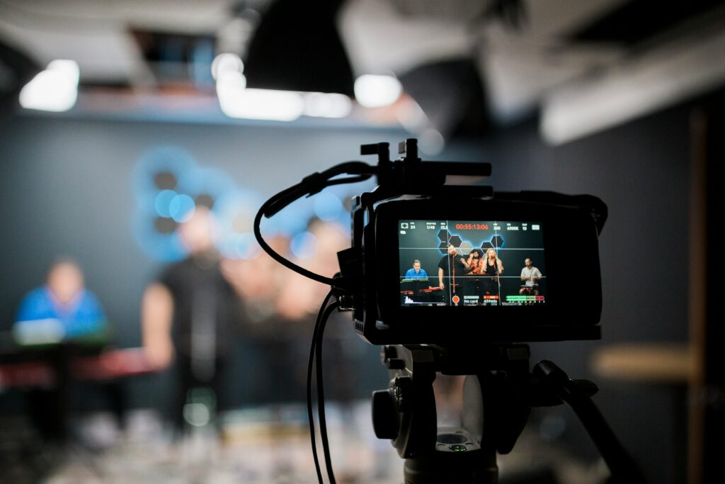 A view through a professional video camera’s viewfinder, showing a group of people performing or speaking on a dimly lit stage with blue, hexagon-shaped lights in the background.