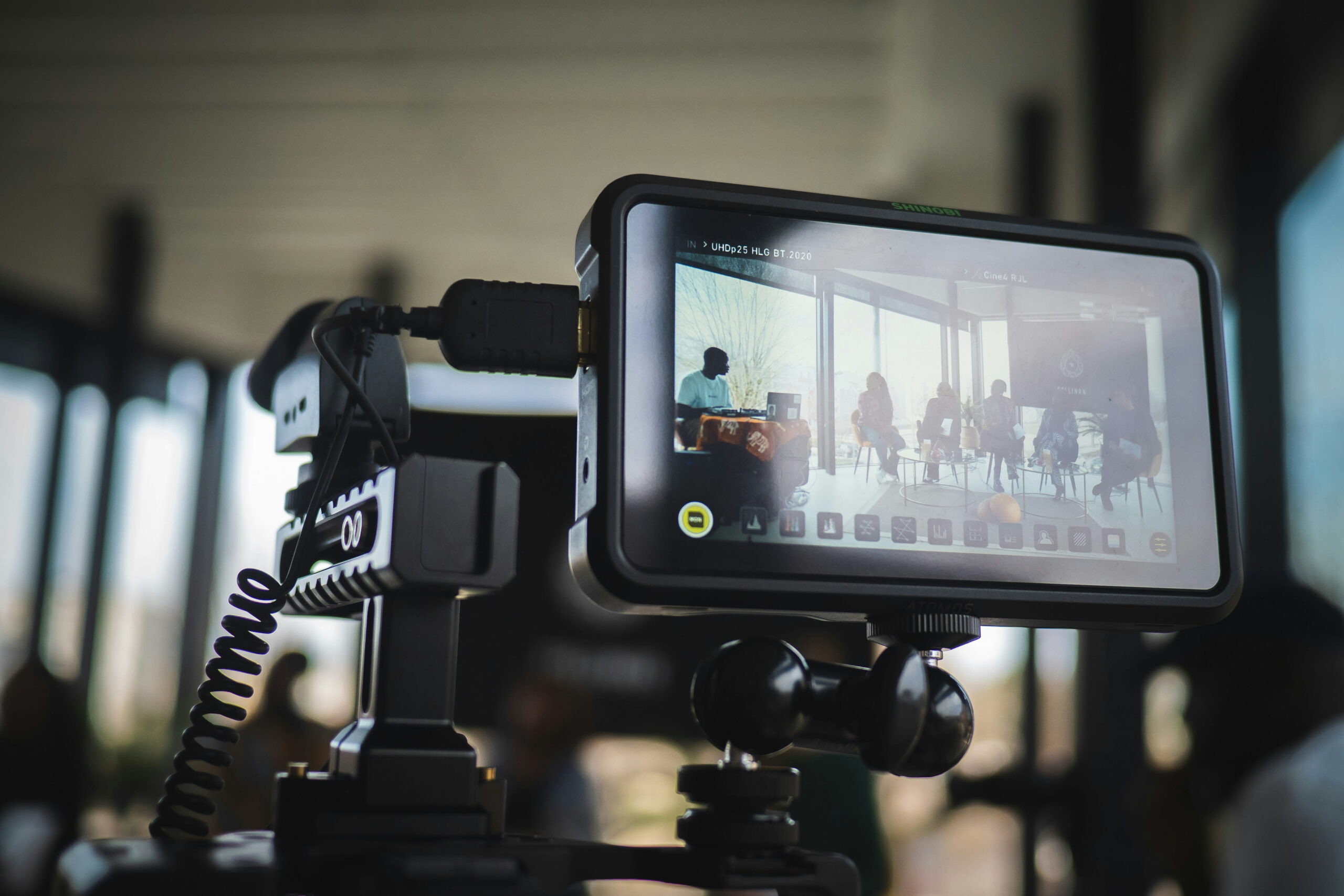 A close-up of an external Atomos Shinobi camera monitor mounted on a rig. The monitor displays a live camera feed of a panel discussion taking place in a bright, modern room with large windows, featuring several participants seated in a semi-circle.