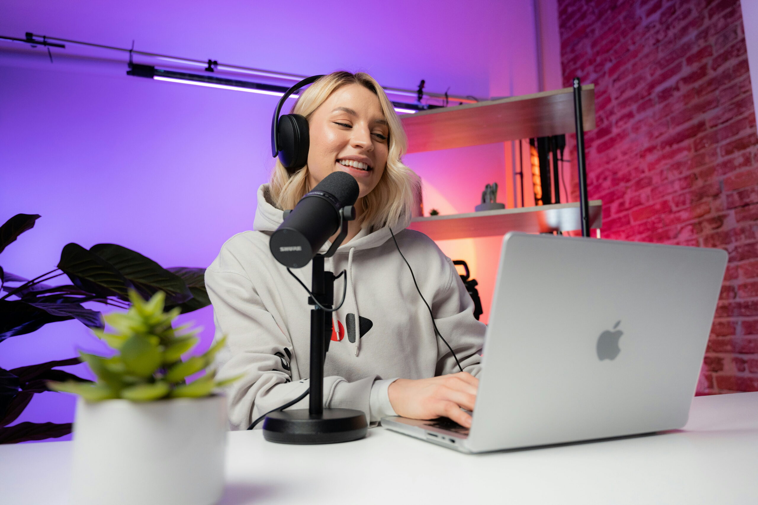 alt text A blond woman wearing headphones and a hoodie sitting at a desk with a laptop and a microphone in a room with purple lighting.