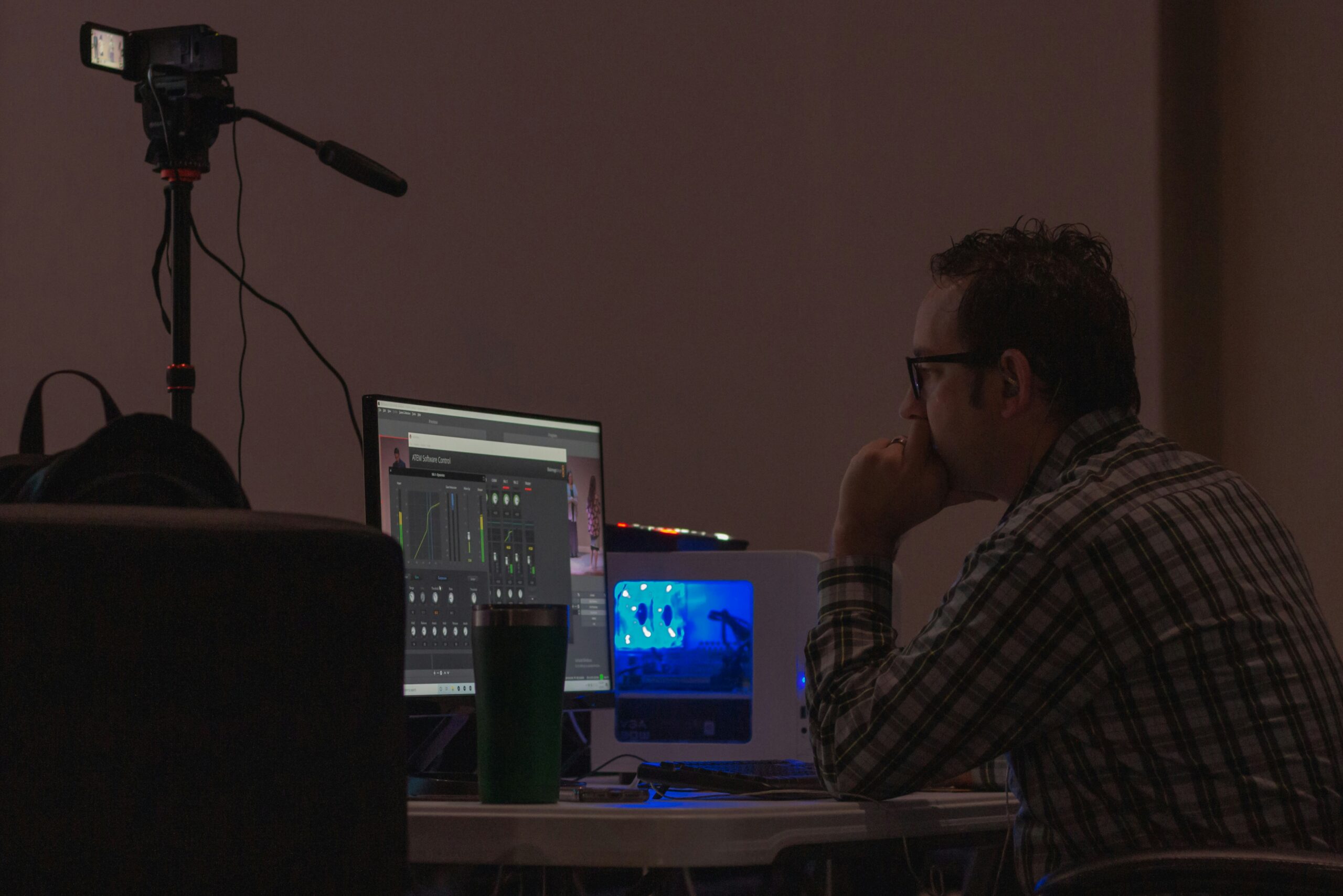 Profile view of a man sitting at a desk with a computer monitor displaying audio control software, a cup, and a video camera on a tripod in the background.