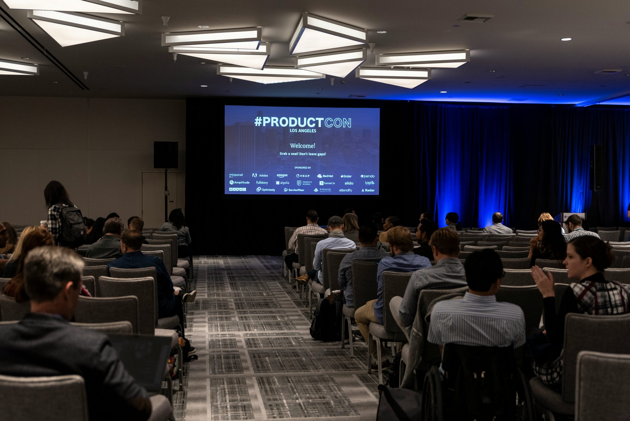 View from the back of an auditorium filled with seated attendees facing a large screen displaying "#ProductCon Los Angeles" with blue stage curtains on the right.