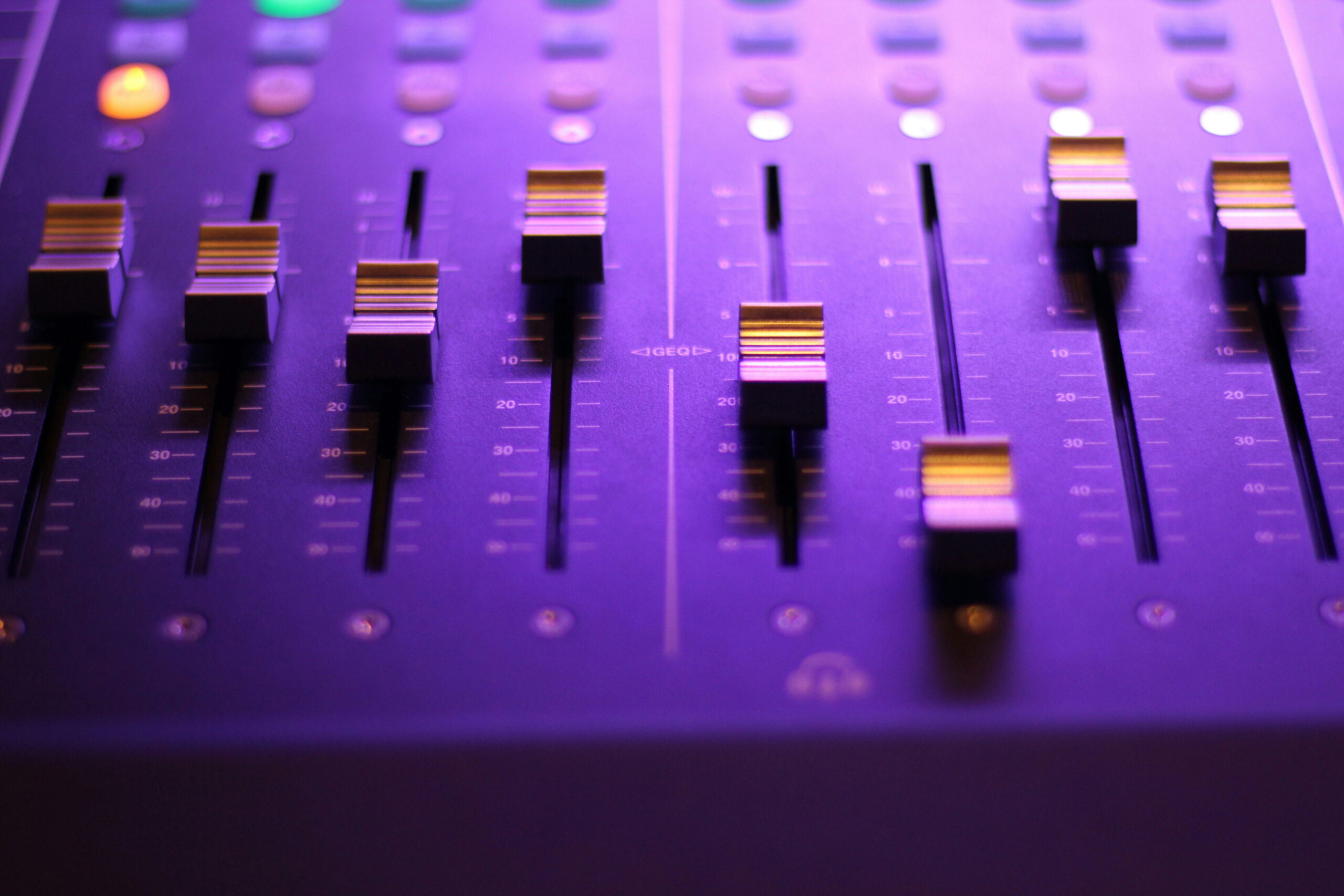 Close-up shot of an audio mixing console illuminated in purple light, focusing on the vertical fader sliders and level markings.