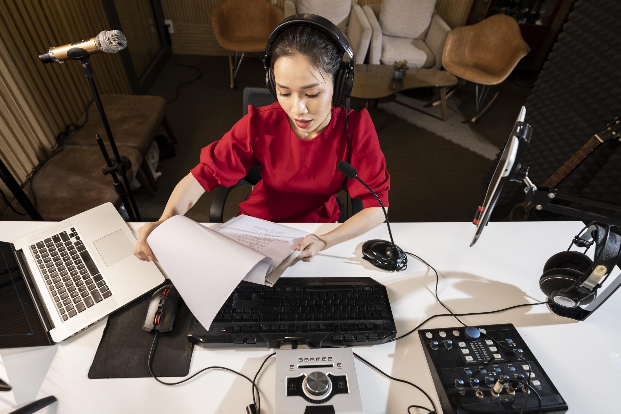 High-angle shot of a woman wearing a red top and headphones, sitting at a white desk while reviewing a script in a studio equipped with microphones, a laptop, and a mixing console.